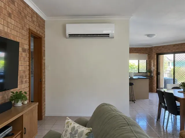 Wall-mounted split system running above a plain living room wall, with open-plan home interior in Nerang